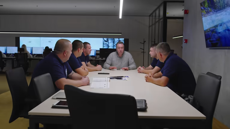 Six men sit around a conference table in a modern office, engaged in a meeting reviewing documents and electronic devices as they discuss a state and local case study.