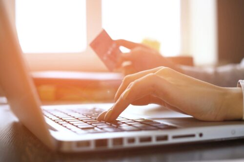 Person typing on a laptop keyboard with one hand while holding a credit card in the other, suggesting online shopping or electronic payment.