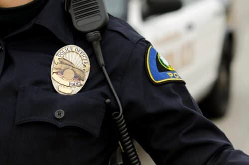 Close-up of a Tribal Nations police officer’s uniform showing a badge, shoulder patch, and radio microphone, with a blurred police vehicle in the background.