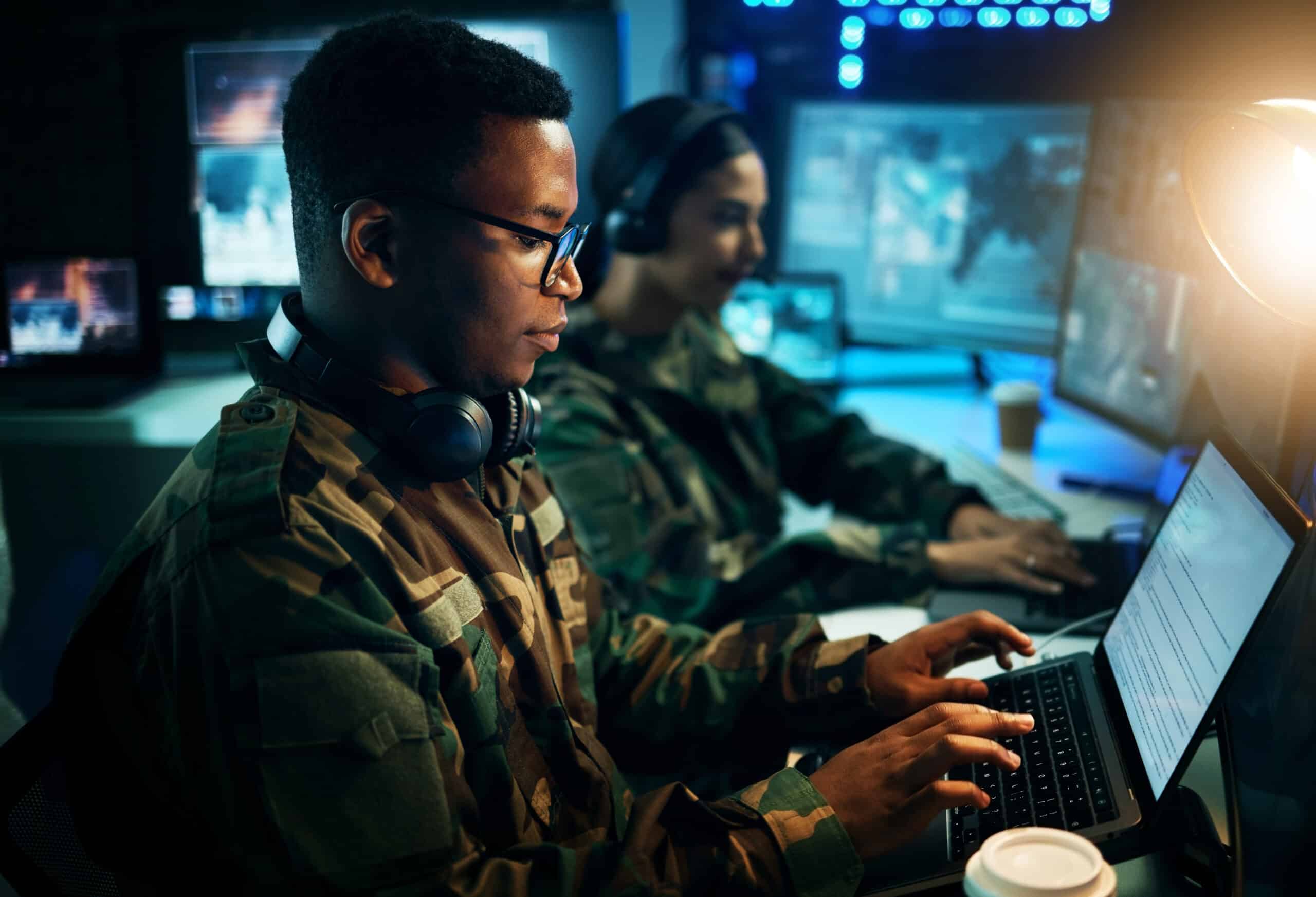 Two military personnel in camouflage uniforms using forensic workstations in a dimly lit control room with multiple screens.
