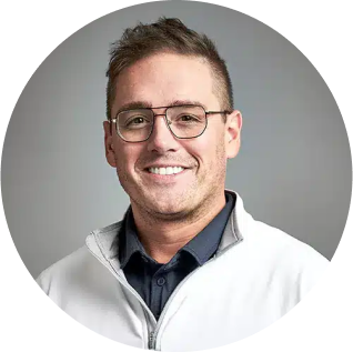 Man wearing glasses and a white jacket, smiling in front of a gray background. Learn more about his new look and confident demeanor.