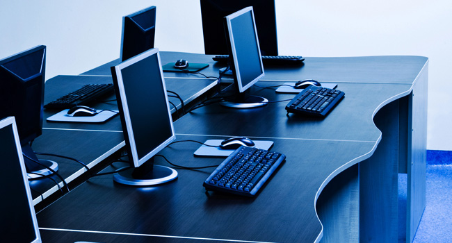 A row of four computer workstations with monitors, keyboards, and mice sits on a dark, curved desk in a blue-tinted room at the new major U.S. research institution dedicated to higher education.